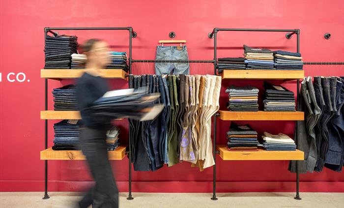 Person walking past shelves of neatly folded denim against a bold red wall at Hiut Denim. Various jeans styles and colours are displayed, with one pai